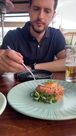 A person, presumably male, is seated at a wooden table and appears to be preparing to eat a dish. He is wearing a dark polo shirt and a wristwatch.  On the table is a light green plate holding a mound of what appears to be tartare surrounded by small green leaves and topped with an orange garnish. To the left of the plate is an empty light green bowl.  To the right is a glass of beer. The person is using a fork to take a piece of the tartare.  Behind the plates is a small black plate and a blurred outdoor background with foliage and buildings. The setting appears to be a patio or outdoor restaurant.
