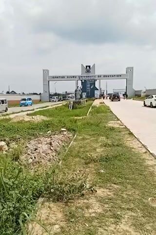 The video shows a gate to Ignatius Ajuru University of Education. Several vehicles, including white and blue vans and a white car, are driving on the road leading to the gate. A person walks along the side of the road. There is a statue in front of the gate. The sky is cloudy. The surrounding area includes grassy patches with some bare soil and a small amount of debris.