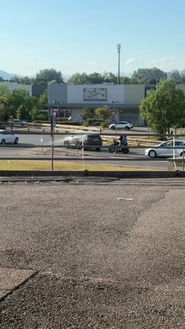 A motorcycle is being ridden by two people. They are riding on a road. There are several cars also on the road. In the background, there is a building with trees and a cell tower. The sky is blue and it appears to be daytime.