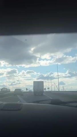 The video captures a view from inside a moving vehicle, looking out through the windshield onto a multi-lane highway during the daytime. The sky is partially cloudy, with patches of bright blue sky visible between fluffy white and grey clouds, suggesting good weather conditions. Sunlight is peeking through the clouds, illuminating the scene.

Ahead on the highway, a large white box truck occupies the middle lane. Several other cars are visible further down the road in various lanes, appearing to be traveling in the same direction. Streetlights and overhead gantries with road signs are spaced along both sides of the highway. Some yellow diamond-shaped signs with black symbols are visible, likely warning signs for drivers. The car's dashboard is visible at the bottom of the frame. The audio includes indistinct talking and general vehicle sounds, typical of being inside a car while driving. No specific readable text is discernible on any signs or vehicles. The setting is Костенеево, Russia.