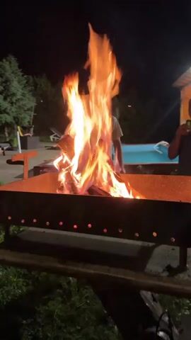 At night, an outdoor scene shows a large fire burning in a metal fire pit. Logs are engulfed in bright orange and yellow flames, with smoke rising. In the foreground, a person's hands hold a green beverage can in their left hand and a fork with a piece of dark-colored food (possibly grilled meat) in their right hand, moving it towards the fire pit. In the background, a young man in a light-colored t-shirt stands near the fire, observing. To the right, a swimming pool is visible, and to the left, dark green trees are present. The only light source appears to be the fire itself.