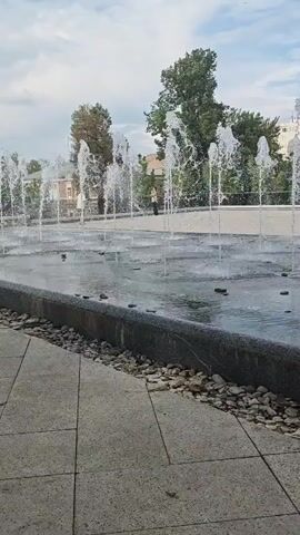 The video shows a shallow, rectangular fountain made of dark gray stone with a border of small gray and white rocks. Multiple jets of water shoot vertically into the air, creating a refreshing display. The water collects in the fountain's basin, creating a reflective surface. In the background, trees and a few buildings are visible under a blue sky with scattered clouds. People can be seen walking and sitting on benches in the park beyond the fountain. The camera is positioned at ground level, offering a low-angle perspective of the fountain and its surroundings.