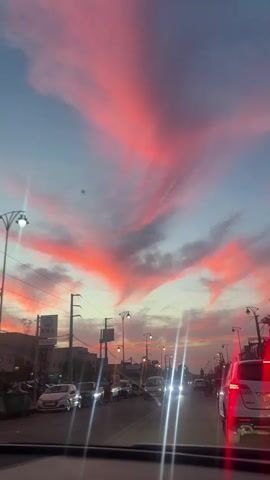 The video shows a car's dashboard view of a street with cars driving during a sunset. The sky is visible and has pink and gray clouds. Buildings and streetlights line the street. The camera is static. The focus is on the sky.