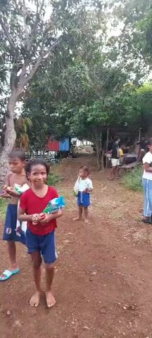 The video shows a group of people, including children and adults, in an outdoor environment. The ground appears to be dirt. There are some structures that look like small buildings or shacks in the background. Trees and vegetation are visible. Some individuals are standing and looking around, while others are walking. The clothing of the people is varied, with some wearing t-shirts and shorts, and others wearing jeans and brightly colored tops. One structure has blue and white painting on it. The overall impression is a casual gathering in a semi-rural or village setting.