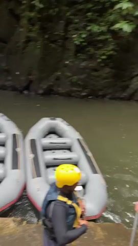 A group of people in inflatable rafts prepare to go whitewater rafting. The rafts are gray with black accents and have seats for multiple people. Some individuals are wearing life vests and yellow helmets. The scene takes place in a lush, green environment with a river flowing between rocky cliffs covered in vegetation.