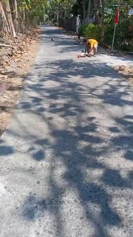 The video shows two children on a paved road under trees. The child in the foreground is wearing a yellow shirt and crouches on a skateboard, holding a small red toy. Another child sits further down the road.