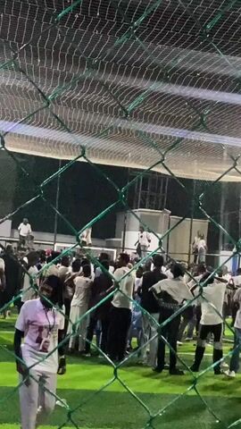 The video shows a crowd of people gathered indoors on a green artificial turf surface, possibly a sports field or event space. The crowd appears to be watching something happening on a raised platform or stage in the background, where several individuals are visible. The scene is viewed through a green chain-link fence, which obscures some of the details. The lighting suggests it is nighttime or an indoor artificial lighting environment. Some individuals in the crowd are wearing white shirts. There are lights illuminating the area, and a net is visible overhead. The overall atmosphere appears to be one of excitement or attention directed towards the elevated area.