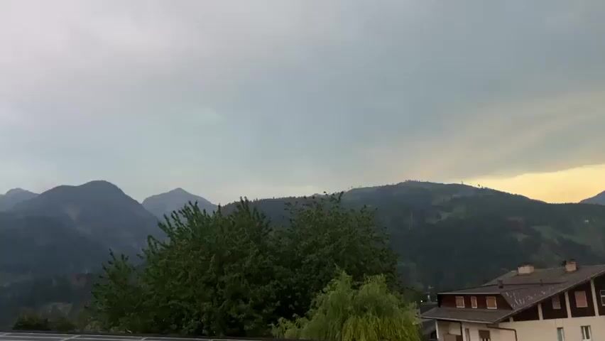 The video pans across a mountainous landscape at dusk. In the foreground, solar panels are visible on a rooftop. Behind the solar panels, several buildings, including a white multi-story building with brown accents and a smaller building with a pitched roof, are nestled amongst trees. A small playground with a swing set is visible in the yard of one of the buildings. The middle ground is dominated by lush green trees and the dark silhouettes of forested mountains. The background features more distant, hazy mountain ranges. The sky is a gradient of grey clouds transitioning to a pale orange and yellow glow near the horizon, indicating the setting sun. No people are visible in the video.