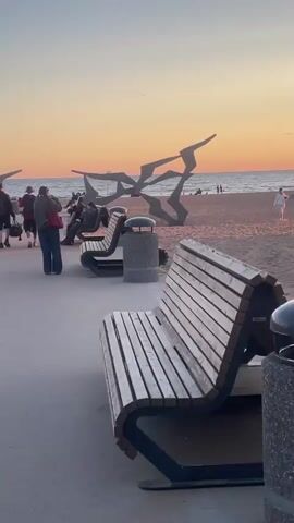 The video shows a beach promenade with several people walking and sitting on benches. The sun is setting, casting a warm orange glow on the sky and the sand. There are benches and trash cans along the promenade, and a sculpture of birds in flight in the background. Some people are walking their dogs, while others are enjoying the view of the ocean. The overall atmosphere is peaceful and relaxing.
