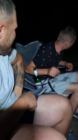 A nighttime outdoor scene shows a person in a black shirt with a pattern of red fruit and gray shorts bending over a table laden with plates of food and cups. Another person, partially visible in the foreground, wearing a gray t-shirt and shorts with a tattoo on their arm, is reaching out towards the bending person. A third person, also partially visible, is seated. A bottle of beer rests in a cup holder attached to a chair. The lighting suggests it is dark outside, with artificial light illuminating the immediate area.