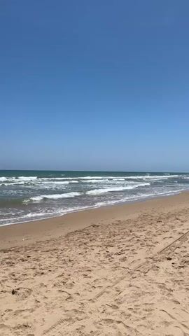 A static shot of a wide beach with the ocean to the left and sand dunes and a fence line to the right under a clear blue sky. The ocean features gentle waves with white caps, and the sandy beach displays numerous footprints and tire tracks.