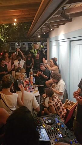 Here's a detailed description of the video:

The video shows a lively party scene in an outdoor venue. A large crowd of people are gathered, with many raising their hands in the air, clapping, and appearing to dance or celebrate. The environment is decorated with string lights overhead, giving it a festive atmosphere. Some of the crowd members are holding drinks. 

In the foreground, a DJ setup with a mixing console is visible. Behind the DJ equipment, a group of musicians is performing. The band consists of a guitarist, a flutist, a trumpet player, a drummer, and what appears to be a banjo player. They are arranged around a table that holds various objects, including drinks, a bottle of water, and other decorative items.

The people in the crowd are diverse in appearance. The overall atmosphere is energetic and celebratory.