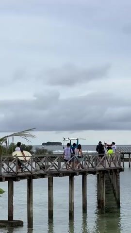 The video shows a wooden boardwalk extending over the water. Several people are on the boardwalk, some of whom are carrying surfboards. One person in the foreground is holding a surfboard upright. The sky is overcast with clouds, and there are waves visible in the distance, with a small island or landmass on the horizon. The water beneath the boardwalk is clear, allowing the wooden pilings to be seen. The scene appears to be a coastal area, possibly a beach or a surf spot, with a calm but cloudy atmosphere.
