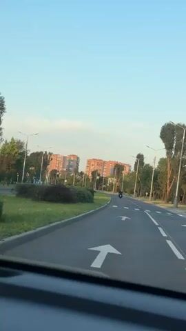 The video shows a road with a motorcycle in the distance. The road has a white arrow painted on it, indicating a turn to the right. There are streetlights and trees lining the road, and in the background, there are tall apartment buildings. The sky is clear and blue.