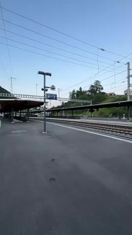 The video shows a train station platform. The platform is paved with asphalt. There are train tracks next to the platform. Overhead, there are electrical wires and poles for the train system. In the background, there are covered waiting areas with benches, buildings, and trees on a hill. The sky is clear and blue, suggesting it is daytime. There are signs at the station, one of which is blue and white and appears to be a track number sign. Another sign, possibly an electronic display board, is visible on a pole further down the platform. The camera appears to be panning from left to right, offering a panoramic view of the station. No people are visible in the video.
