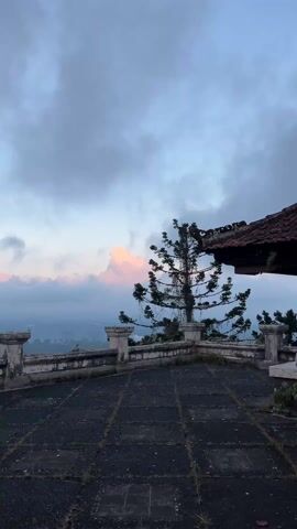 The video shows a view from an abandoned structure, likely a balcony or rooftop, with a stone tile floor and a decaying stone railing. A small, sparsely needled tree stands near the railing. Beyond the railing, a landscape of hills or mountains is visible, shrouded in mist or fog. The sky is overcast with grey clouds, but a patch of pink-tinged clouds suggests the possibility of a sunrise or sunset. A portion of the structure's roof, made of red tiles, is visible in the upper right corner of the frame, appearing weathered and overgrown with vegetation. The overall atmosphere is quiet and somewhat melancholic.