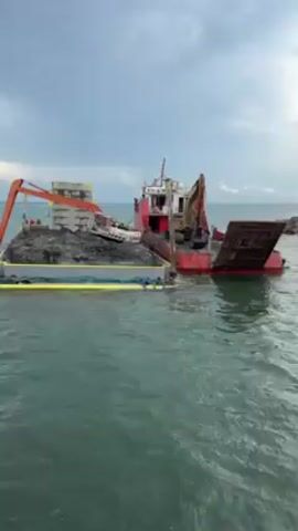 The video shows a red and white barge in a body of water near a shoreline. The barge is loaded with dark, gravel-like material, some concrete blocks, and a white vehicle on top. Two excavators are visible on the barge, one orange and one yellow. A yellow containment boom surrounds the load. The water is slightly choppy. The sky is overcast. People can be seen working near the orange excavator. The barge appears to be carrying materials to the shore.