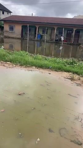 The video shows a flooded street in front of a building. The building appears to be a residential structure with multiple windows and a covered porch area. Some people are visible in front of the building, and there are water barrels and other objects on the porch. The water level is high enough to flood the lower part of the building and create reflections of the structure in the water. The street in the foreground is also flooded and appears to be unpaved, with some debris scattered in the water. The overall scene suggests a recent heavy rainfall or a drainage issue.