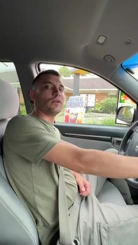 A man with short brown hair and a beard, wearing a green t-shirt and light-colored pants, is sitting in the driver's seat of a car. He is looking at the camera and smiling. A black dog is sitting in the passenger seat, looking towards the man. The car appears to be at a drive-thru window, with a menu board visible outside the car. The man's left arm is resting on the steering wheel, and his seatbelt is buckled. The interior of the car is visible, including the dashboard and the car's ceiling. The dog has a pink tongue sticking out of its mouth.