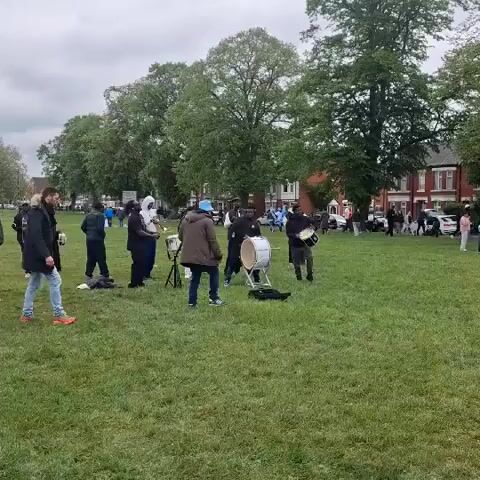 The scene captures a vibrant outdoor gathering on a green field or park. In the center, a small band of musicians is playing. The band includes individuals with a trombone, drums, and other instruments. Several people are standing around watching or listening to the performance. In the background, more people are visible, suggesting a larger event or gathering. The setting is somewhat urban, with houses and cars visible beyond the trees. The weather appears overcast, casting a subdued light over the scene.