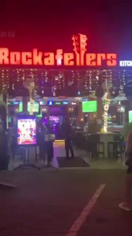 The video shows the exterior entrance of a bar and restaurant named "Kateller's Kitchen" at night. The establishment is brightly lit, with a red neon sign and festive lights. Several flat-screen televisions are visible, some displaying what appears to be a sporting event. Inside, there are people seated at tables and a band performing on a small stage with a blue and white light display. A doorman in dark clothing stands near the entrance. A woman with long blonde hair walks into the bar.