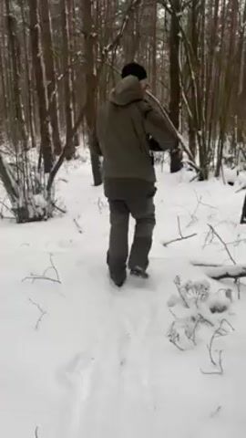 A person is walking through a snowy forest. The individual is wearing a green jacket, grey pants, a black hat, and is carrying what appears to be a long object over their shoulder. The camera follows them from behind. The forest consists of tall, bare trees and a ground covered in snow. Other people are visible in the background. The snow appears to have a light, powdery texture.