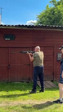 A man is standing in a grassy area in front of a wooden shed, holding a rifle. He is aiming the rifle towards the right side of the frame. The man is wearing a camouflage t-shirt, dark pants, and black boots. The shed is made of dark red wood and has two large doors. There is a small oval window on the left side of the shed. A padlock is visible on the left door. The sky is blue with some clouds, and the sun is shining, suggesting it is daytime. Green grass covers the ground, and some trees are visible behind the shed. A partial view of another person wearing blue shorts and black and white sneakers is visible on the far right of the frame. The scene appears to be outdoors in a rural or suburban setting. There is text visible on a small sign mounted on the right side of the shed, appearing to be a house number, but it is too blurry to read definitively. The overall mood appears to be casual, possibly during a recreational activity like target practice.