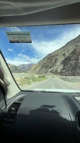 The video shows a view from inside a vehicle, looking out through the windshield and side window. The scene depicts a mountainous landscape with a winding road. The mountains are dry and appear to be made of rock and soil, with patches of vegetation. The sky is blue with white clouds. A small yellow building with "BORDER ROADS ORGANISATIO" written on it is visible on the side of the road. The camera is moving, suggesting the vehicle is in motion. There are no people visible in the video. The interior of the vehicle is partially visible, including the dashboard and a side mirror. A sticker is affixed to the windshield.