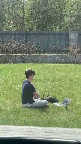 A medium shot depicts a young person sitting cross-legged on green grass in a backyard, next to a laptop and a black dachshund. The person wears a dark blue t-shirt and light-colored pants, and appears to be looking at the laptop screen. The dachshund is lying on its back next to the person. A grey fence is in the background with sparse bushes and trees behind it. The sun is shining and casting shadows on the grass. Someone is audibly laughing.