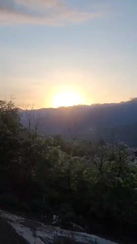 The video captures a scenic mountain landscape at sunset. The sun is partially obscured by a mountain ridge, casting a warm glow across the sky and the surrounding trees. The sky is a soft blend of blue and yellow, with a few wispy clouds. Dense foliage, characterized by lush green trees and some bare branches, dominates the foreground and midground. The terrain appears to be uneven, with a dirt path visible in the lower left corner. In the far distance, a few buildings suggest a settlement nestled within the mountains. The overall atmosphere is serene and tranquil, evoking a sense of peace and natural beauty. The time of day is clearly indicated as sunset due to the low position of the sun and the warm, diffused lighting. The setting is a mountainous region, with the presence of trees and hills suggesting a rural or natural environment. No text is visible in the video.
