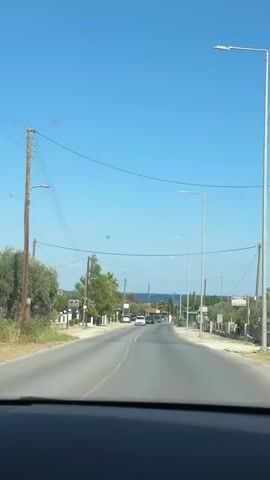 The video shows a view from inside a vehicle driving down a paved road in a rural or coastal area. The road is lined with trees, some of which appear to be olive trees, and buildings on either side. There are several cars on the road, and power lines are visible overhead. The sky is clear and blue, and in the distance, the sea is visible. The sun appears to be shining brightly. The video captures a sense of movement and travel, as the scenery slowly passes by.