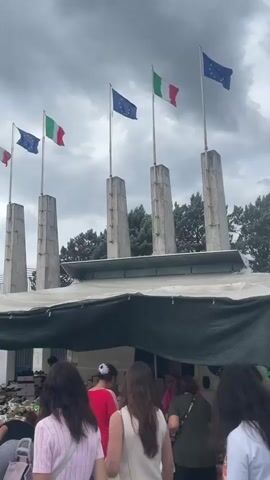 The video shows a crowded outdoor market on a cloudy day. Several Italian and European Union flags fly from poles above a row of stalls. People are browsing merchandise, which appears to include clothing and other goods. The overall atmosphere is busy and somewhat chaotic, with many individuals walking and interacting.