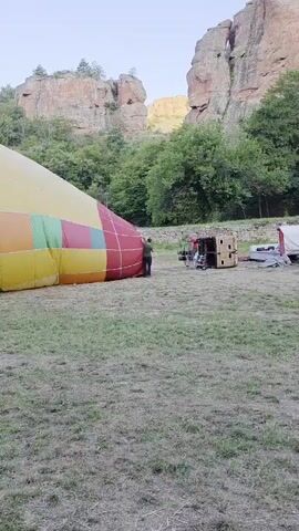 A hot air balloon, predominantly yellow with a band of red, orange, green, and yellow stripes around its base, lies partially deflated on a grassy field. A person in a dark shirt is standing near the opening of the balloon, appearing to be involved in its preparation. In the background, a tan-colored basket, likely for the hot air balloon, rests on the ground next to some equipment. Behind the field, a rugged, rocky cliff face rises, with trees and foliage growing on its slopes. The sky above is overcast and light.