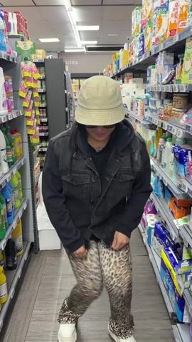 A person dances in a supermarket aisle. The individual wears sunglasses, a bucket hat, a black hoodie under a denim jacket, and leopard-print pants with matching shoes. The person performs dance moves, including squats and hand gestures. The background shows shelves stocked with various household and cleaning products. The aisle is narrow and brightly lit.