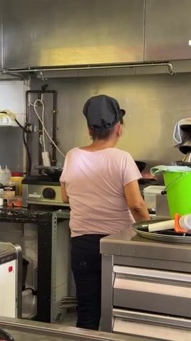 A woman wearing a pink t-shirt, black pants, and a black hair covering, and black gloves, stands with her back to the camera in a commercial kitchen. She is facing a stove where a wok is heating with flames visible beneath it. To her left, on the counter, are various bottles and containers. To her right, a bright green bucket is visible. In the foreground, a red counter separates the viewer from the kitchen area. Another person, a woman with dark hair, is partially visible in the lower left corner, facing away from the camera. The kitchen environment features stainless steel surfaces, shelving with bottles, and a yellow-painted wall.