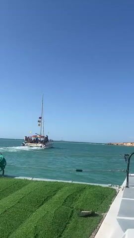 The video shows a boat sailing in the ocean on a clear, sunny day. The camera appears to be on another boat, with a small patch of artificial turf and a metal railing visible in the foreground. A person's legs and bare feet are visible in the lower center of the frame. In the distance, a boat full of people is moving away from the camera. A red and white lighthouse stands on a rocky jetty in the background. The ocean is a clear turquoise color.