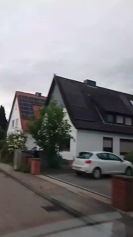 A video pans across a residential street. On the left, a white house with a red tiled roof features solar panels. Further down the street, a darker house with a gray slate roof also has solar panels. Several cars are parked along the street, including a white vehicle, a black hatchback, and a dark-colored SUV. Green trees and hedges line the properties. The sky is overcast.