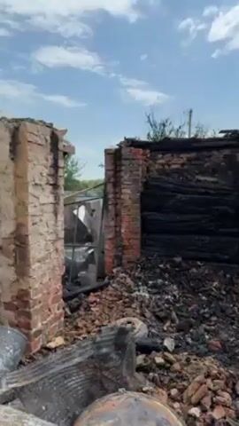 The video shows a burned-out building. The walls are partially standing, made of red brick. The interior is filled with charred wood and debris. The sky is visible above, with scattered clouds. There are remnants of household items amidst the rubble. There is no one visible in the video.