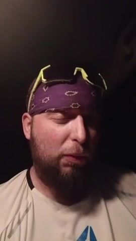 A close-up shot shows a man wearing a purple bandana with white paisley designs and yellow sunglasses pushed back on his head. He has a beard and is wearing a white shirt with a black design on the left side. He is speaking directly to the camera, his expression conveying emotional distress. The background is dark, suggesting an indoor setting with limited lighting.