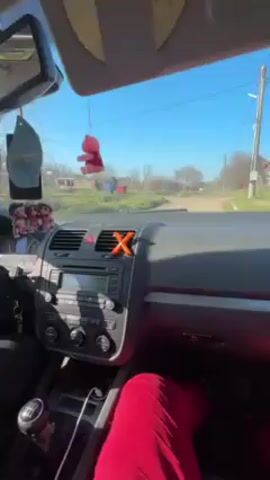 From the interior of a car driving down a country road on a sunny day, a person wearing a black top and red pants manipulates the car radio while loud music plays. The car's dashboard is visible, adorned with an air freshener, a small toy, and an orange X-shaped object. The car has a manual transmission. Beyond the windshield, the landscape consists of a dirt road, green fields, and bare trees under a clear blue sky.