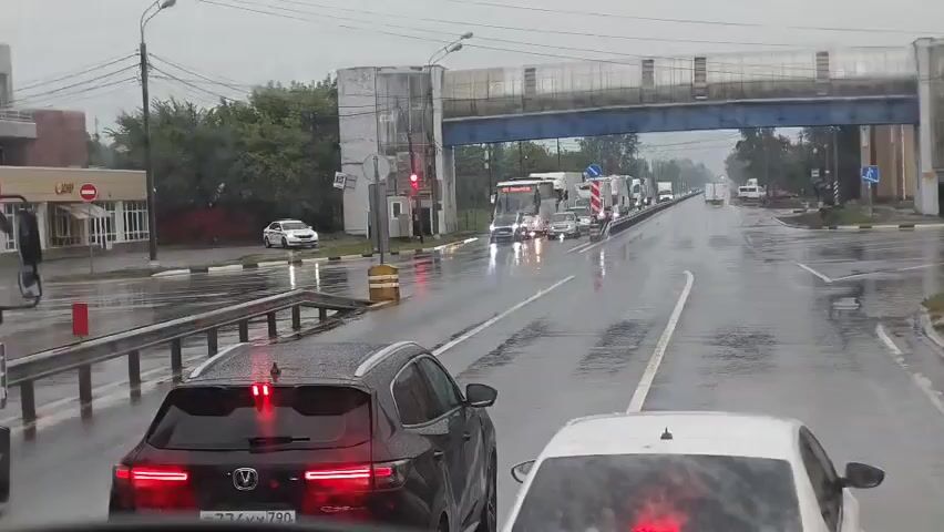 A traffic jam is occurring on a wet, multi-lane road during daylight hours. It appears to be raining, as evidenced by the wet asphalt and water droplets on the windshield. The vehicles are stopped, with brake lights illuminated on several cars in the foreground. A police car with flashing blue and red lights is visible in the distance on the left side of the road. Several large trucks and buses are also part of the traffic congestion. An elevated pedestrian walkway or bridge crosses over the road. The sky is overcast.