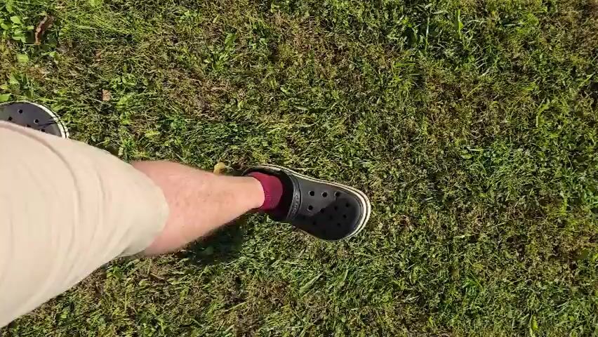 The video shows a first-person perspective of someone walking on grass. The person is wearing beige shorts and black Crocs with red socks. The grass is green with some patches of brown. The camera angle is looking down at the person's feet and the ground. The person takes several steps, with the feet moving from the bottom of the frame towards the top and then back down. The lighting suggests it is daytime with sunlight.