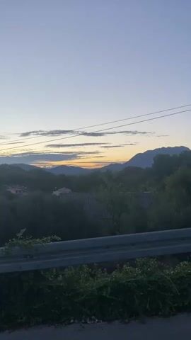 This is a vertical, low-angle, medium shot video taken during either sunrise or sunset. The foreground features a grey metal guardrail that spans the width of the frame, with lush green ivy growing over it from below. Beyond the guardrail, the scene opens up to a landscape of rolling green hills and trees. A few houses with red and grey roofs are visible nestled among the trees. In the distance, a range of mountains forms the horizon, with the sky above displaying soft hues of blue and orange, indicating the time of day. The overall atmosphere is tranquil and serene. There are no people or animals visible in the video. The video consists of a series of still frames, creating a slight stuttering effect as the scene progresses.
