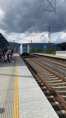 A blue and white train approaches a station platform where several people are waiting. The train has a blue front with a white stripe and the Czech Railways logo. The platform is paved with gray tiles and has a yellow tactile warning strip along the edge. The train stops beside the platform. The sky is cloudy.