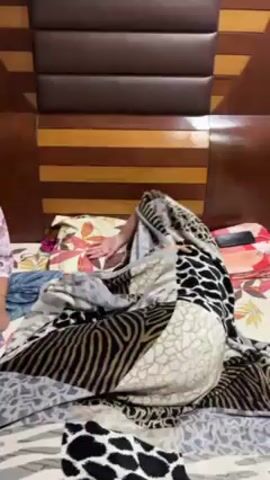 A medium shot shows a young girl on a bed talking towards the camera. She wears glasses, a light floral shirt, and blue pants. A person covered in a black and white patterned blanket is on the bed. The bed has a striped headboard. A pillow with a floral pattern is behind the blanketed person.