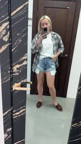 A blonde woman with blonde hair is taking a mirror selfie. She is wearing a white t-shirt with a graphic design, denim shorts, an open grey and white checkered shirt, and brown slip-on shoes. She is standing in what appears to be a room with a large mirror in the foreground, a wooden door in the background, and a dark patterned frame around the mirror. Her left hand is in her pocket, and she is holding her phone in her right hand to take the picture. The overall lighting is bright.