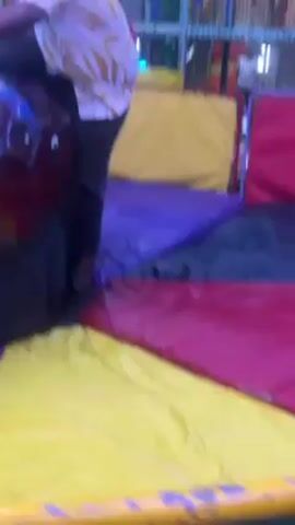 A man is seen from behind, attempting to ride a mechanical bull in what appears to be an indoor amusement park. He is wearing a white shirt with a yellow leaf pattern, dark gray pants, and no shoes. He is holding on to the bull with both hands. The bull is dark red with some blue accents. Around the bull is a multicolored inflatable surface consisting of yellow, purple, red, and green sections.  Other people are standing around watching and recording him, including a man in a red shirt. The amusement park features a climbing structure in the background. The scene is brightly lit.