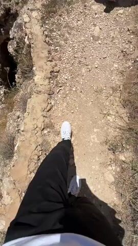 The video depicts a first-person perspective of someone walking on a rocky path, descending towards a valley with a village in the distance. The person filming is wearing black pants and white sneakers. Ahead of them, two other people are also walking down the path. The person in the lead is wearing a gray T-shirt with a design on the back and khaki pants. The sky is blue with a few scattered clouds. The landscape is dry and rocky with sparse vegetation. A simple metal fence can be seen to the right.