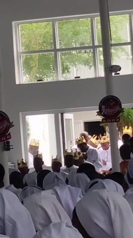Inside a brightly lit building with white walls and large windows, a large group of people are gathered, many wearing white robes. The scene appears to be a ceremonial or religious event taking place in Anantigha, Nigeria.

The foreground shows the backs of several individuals dressed in white, some of whom have their heads bowed, suggesting prayer or reverence. Beyond them, numerous other participants are visible, predominantly men, also clad in white attire. A distinctive feature is that many of these individuals are wearing ornate golden crowns, indicating a significant and perhaps monarchical or spiritual leadership role within the group.

Through open glass doors in the background, more robed figures can be seen, some also wearing crowns. Natural light streams in from the windows, illuminating the space and showing glimpses of green foliage outside, suggesting it is daytime.

Two circular banners are prominently displayed on columns or walls. Each banner features text that includes "THE UNIVERSAL MONARCH" and the year "2006," possibly indicating the founding year or a significant anniversary for the organization. The overall atmosphere conveyed is one of solemnity and devotion, with participants engaged in a communal activity, possibly singing or chanting, given some open mouths visible in the frame.
