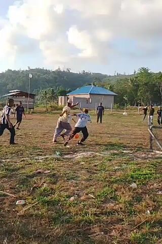The video shows a group of people playing soccer in an open, grassy field. The field appears to be somewhat uneven and dry, with sparse vegetation. In the background, there are trees, a few buildings, and a hill. The sky is partly cloudy. A makeshift goal, constructed from wood, is visible on the field. Several individuals are actively engaged in the game, with some running, kicking a ball, and jumping. The focus is on the gameplay and the interactions of the players. The atmosphere appears casual and recreational.